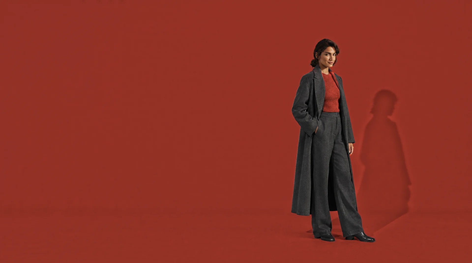 Vrouw met donker haar in een lange antraciet jas en broek met een diep rode top, in een studio die helemaal diep rood is.