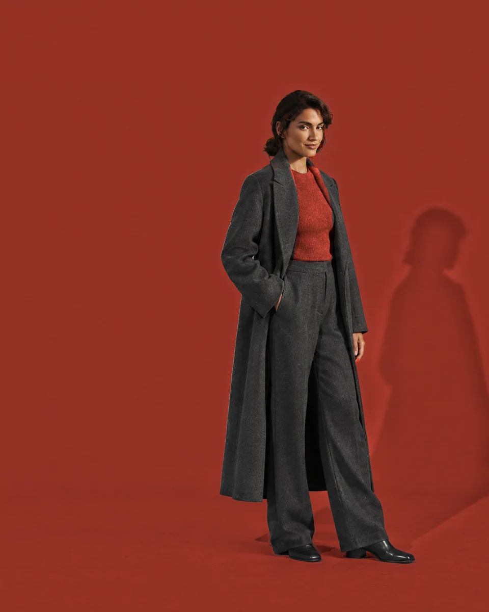 Vrouw met donker haar in een lange antraciet jas en broek met een diep rode top, in een studio die helemaal diep rood is.